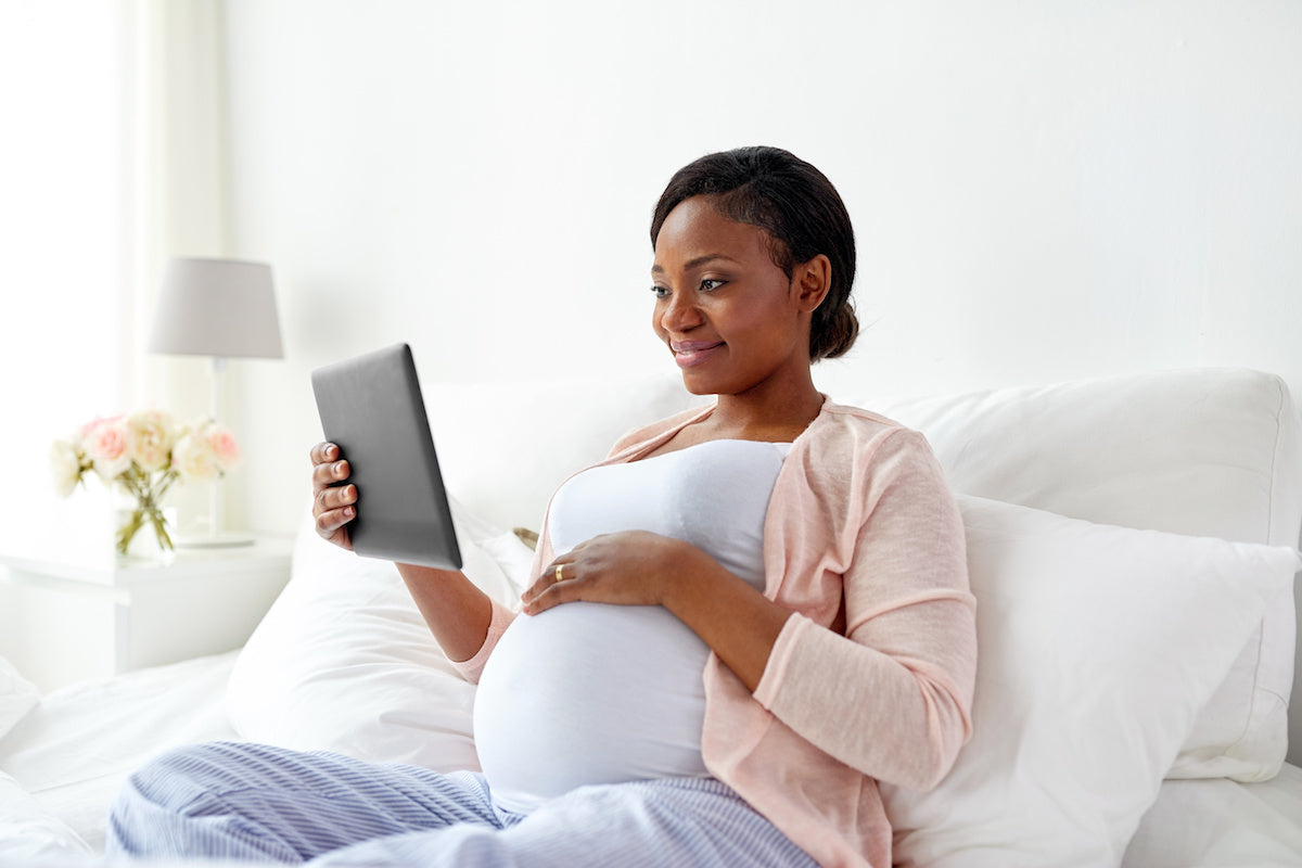 pregnant black mother dressed n pastels sits on a bed reading a tablet
