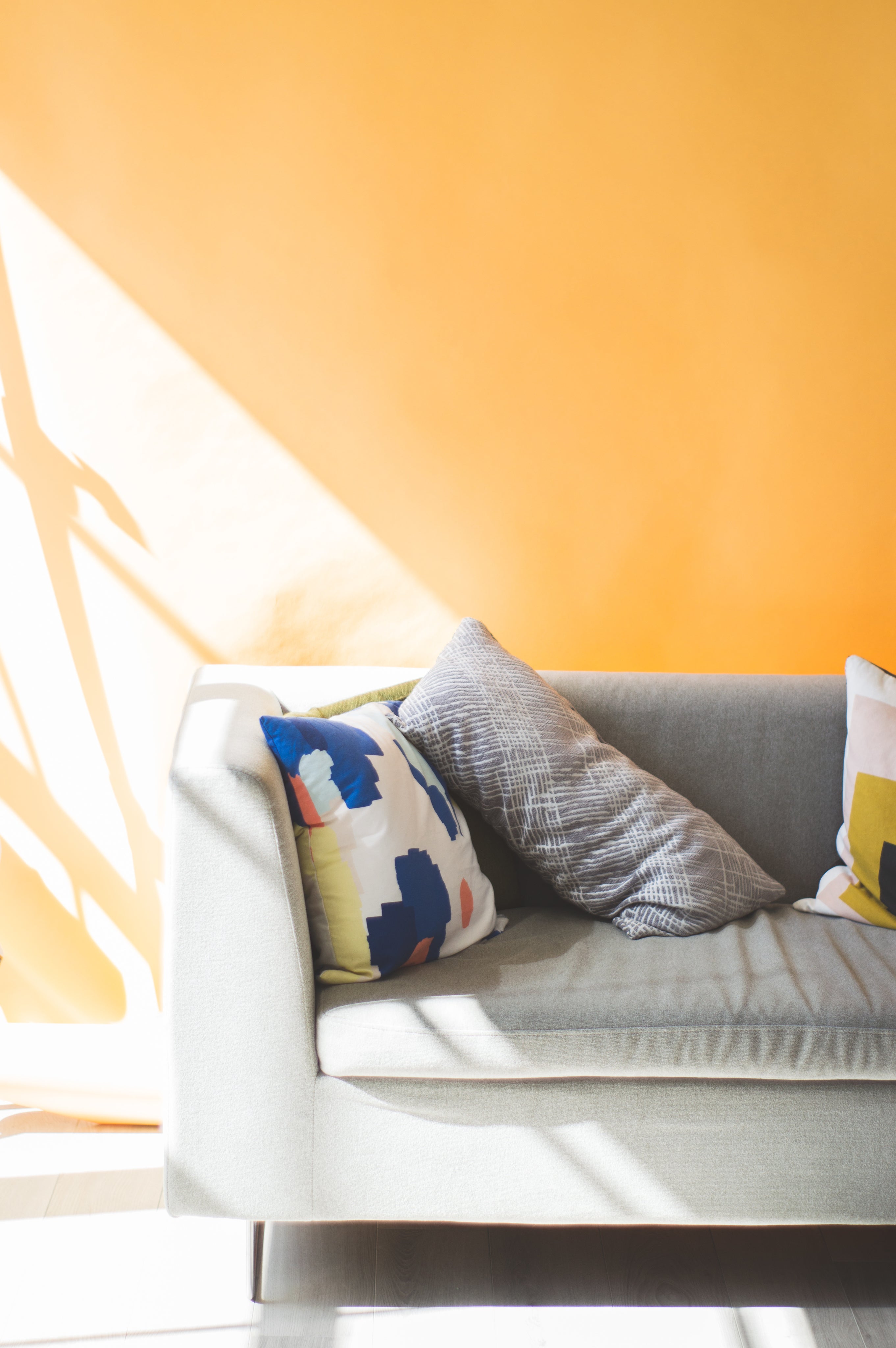 Sun streaming over a grey sofa with multicolor print and grey pillows against a yellow wall