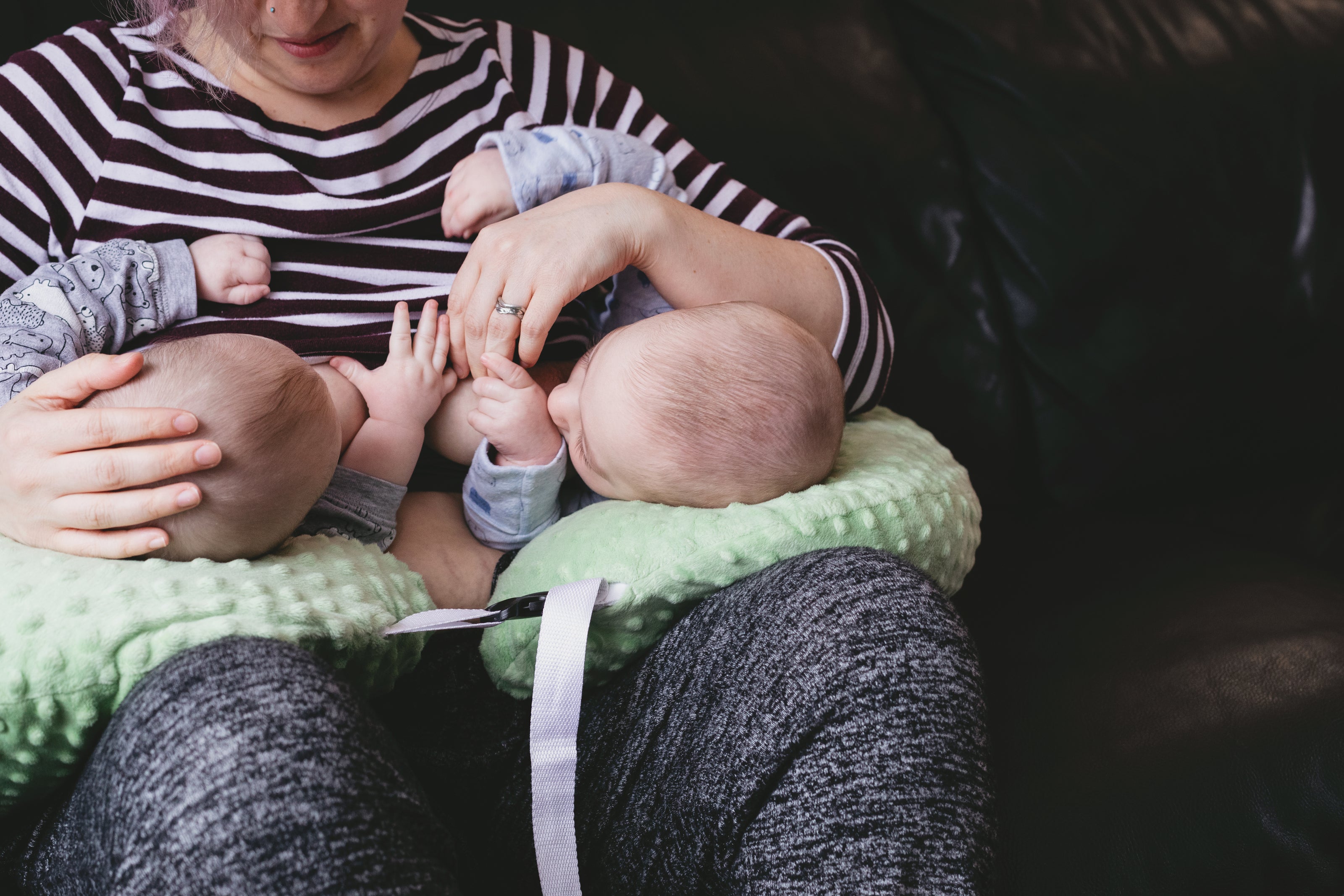 mother in a striped shirt breastfeeding twins who are supported on a light green pillow. Everyone is looking at each other